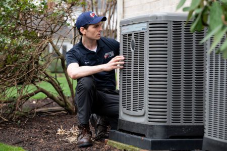 HVAC technician servicing an outdoor HVAC unit next to a home. Bushes in background.